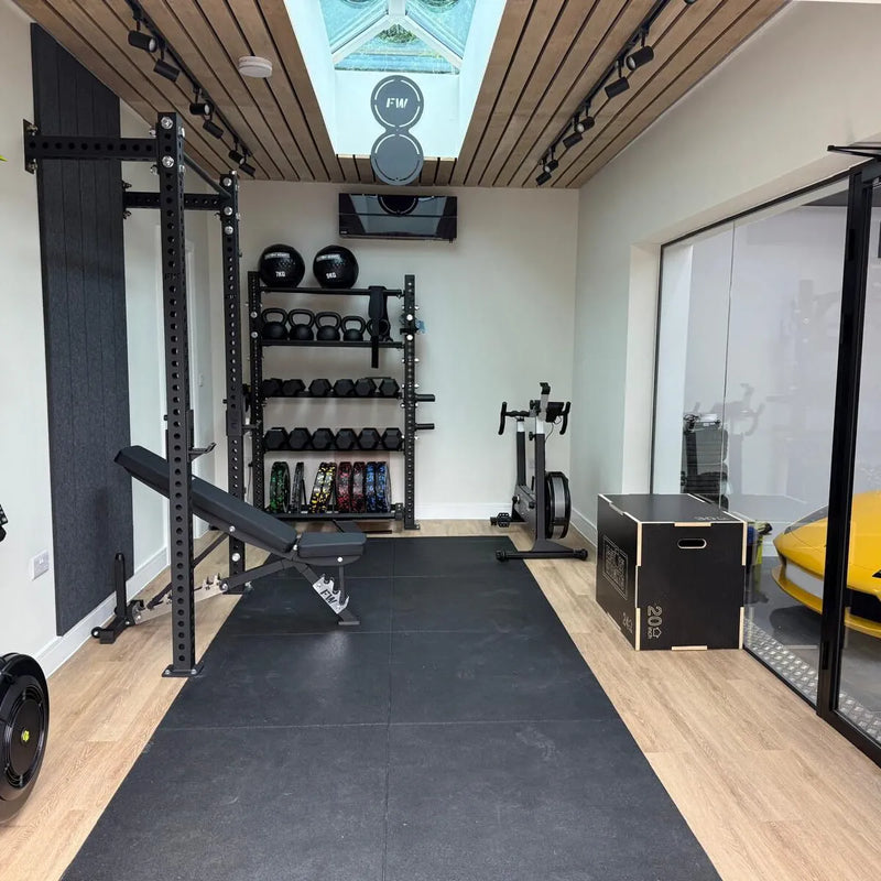 A home gym interior with a black power rack, weight bench, kettlebell rack, exercise bike, plyo box, and black rubber floor mats on light wood flooring. 