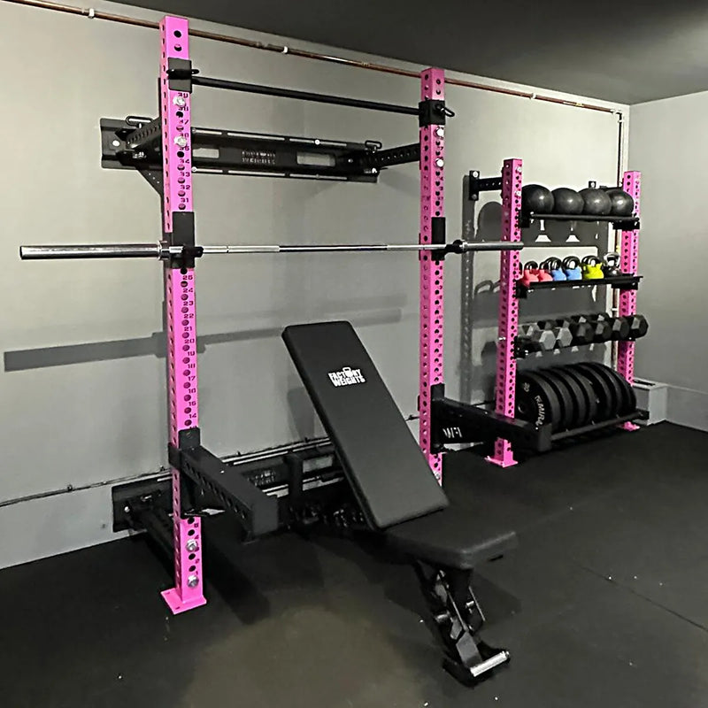 A home gym setup featuring a pink and black power rack with a bench press, a weight bench, and a rack of dumbbells and kettlebells against a gray wall.
