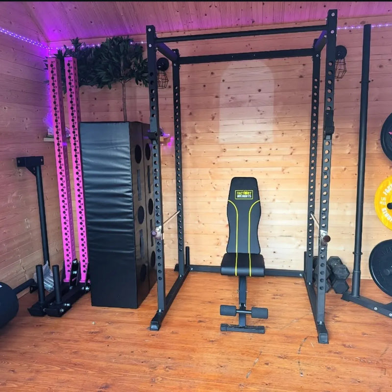 A home gym setup featuring a black power rack with a weight bench inside, a large black plyo box, and weight plates on a wooden wall and floor.