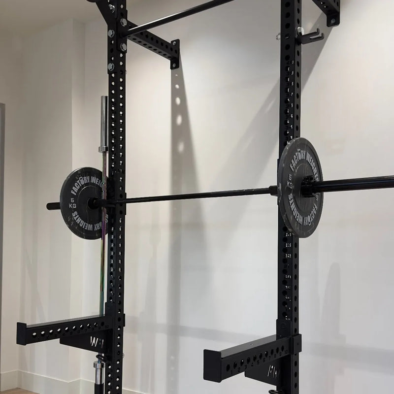 A black power rack with a loaded barbell resting horizontally across the front, photographed in a real indoor room with white walls and baseboards.