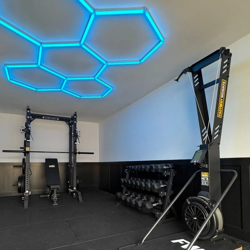 A home gym interior featuring a black adjustable power rack with a barbell, a weight bench, a dumbbell rack, and a large black leg extension machine in the foreground.