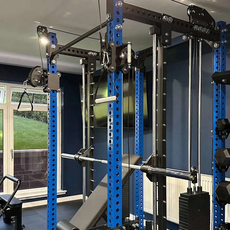 A large home gym power rack system with blue and black steel components, including a pull-up bar, cable pulley system, weight bench, and barbell rack, shown in a home gym setting.