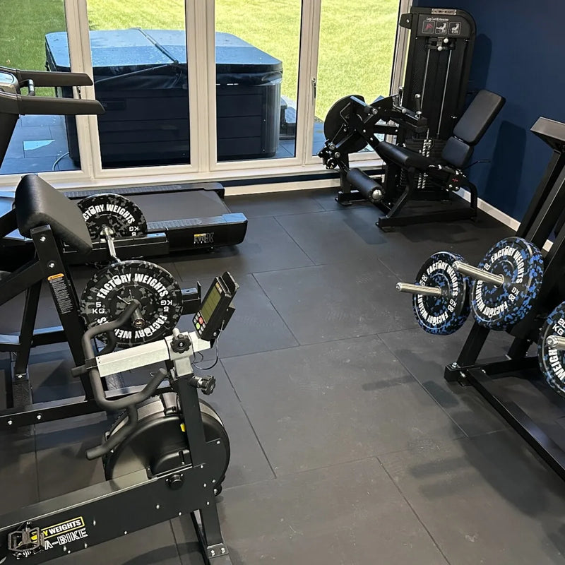 A home gym interior showing multiple weight machines and barbells, including a leg extension in the foreground, a cable machine, and a weight bench near large windows.