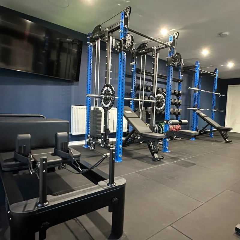 A home gym interior featuring a blue and black power rack with weight plates, a bench, a cable machine, and Pilates reformer machines on a dark tiled floor.
