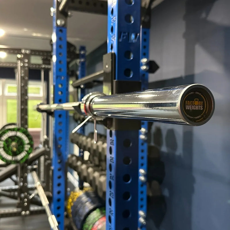 A chrome barbell with a black end cap printed with the Faceay Weights logo, resting horizontally on a blue power rack inside a gym.