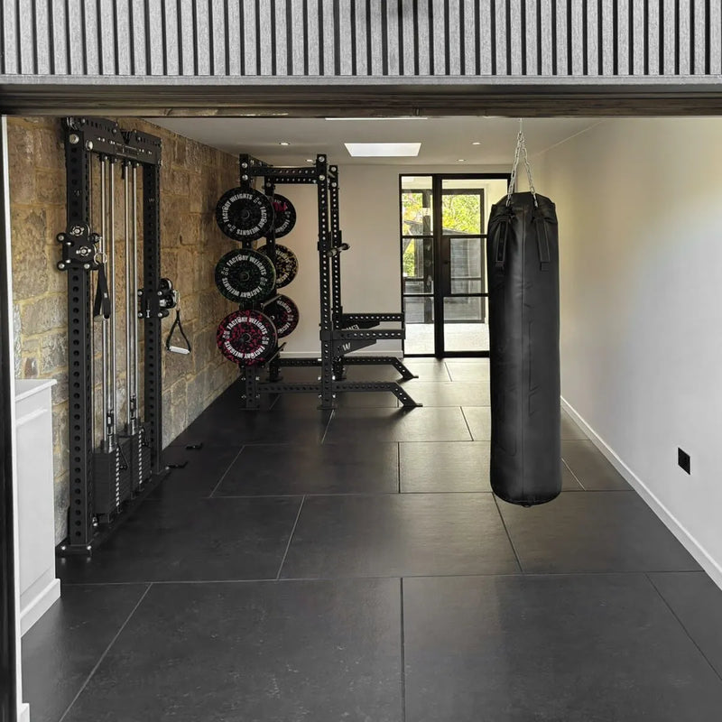 A home gym interior with a black hanging punching bag on the right, a black weight rack with stacked weight plates on the left, and a stone wall behind the equipment. 