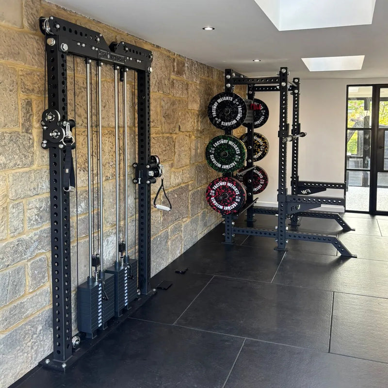 A black cable pulley gym machine mounted on a stone wall, with a dark weight rack and colorful weight plates in the background of a home gym interior.
